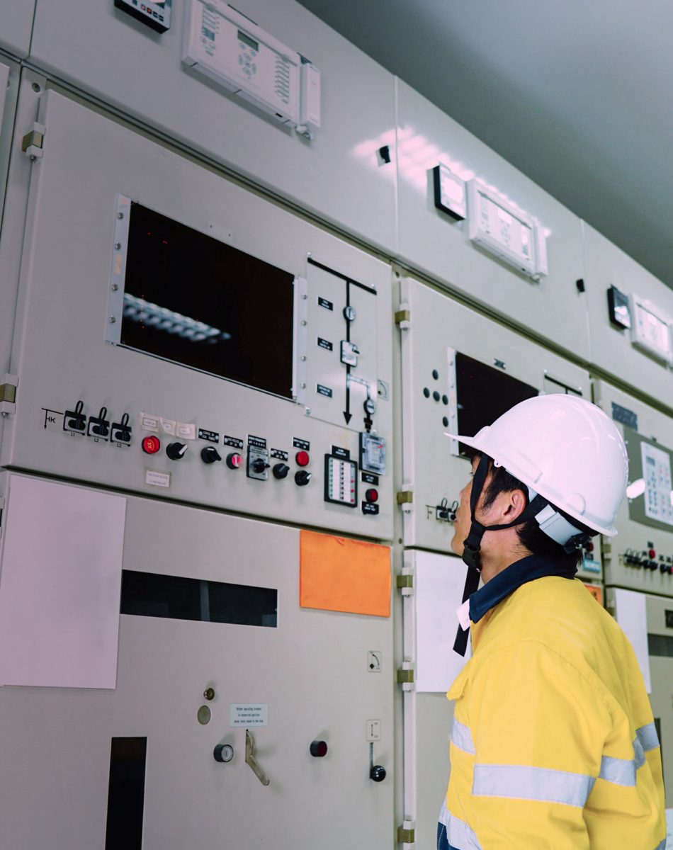 Male engineer wearing a yellow uniform and wearing a white safety hat, inspecting electrical systems in a large power plant.